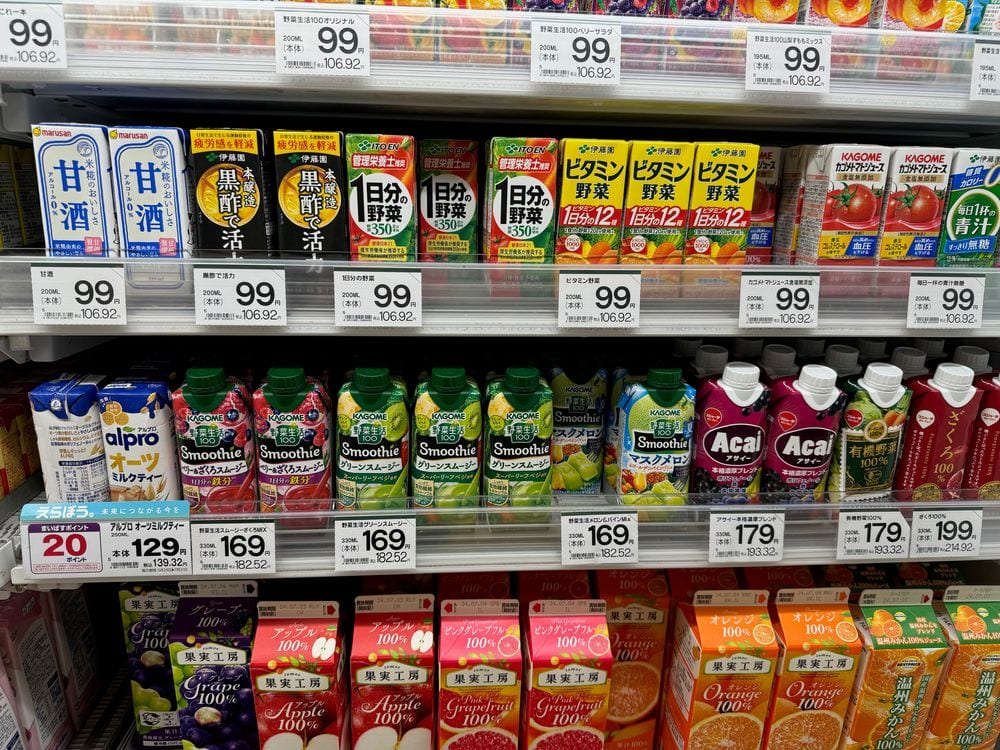 A cold-drink shelf at a Japanese supermarket in Shibamata, Tokyo, with rows of juice, smoothies, and amazake bottles under yellow price tags reading 99, 169, 179, and 199 yen.