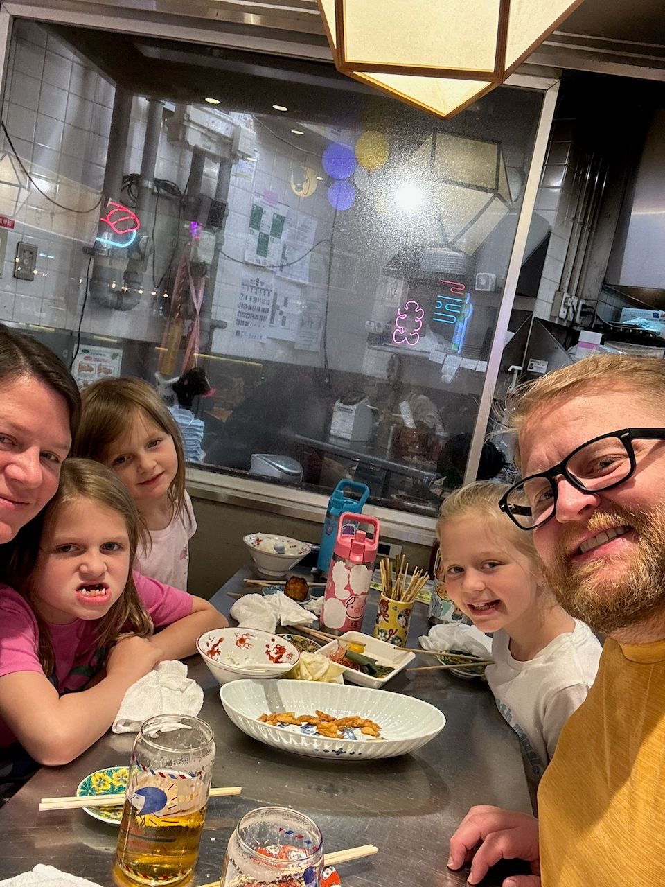 A family selfie at a restaurant table in Asakusa, with two adults and three young girls surrounded by Japanese dishes, chopsticks, and beer glasses, with a view into the open kitchen behind them.