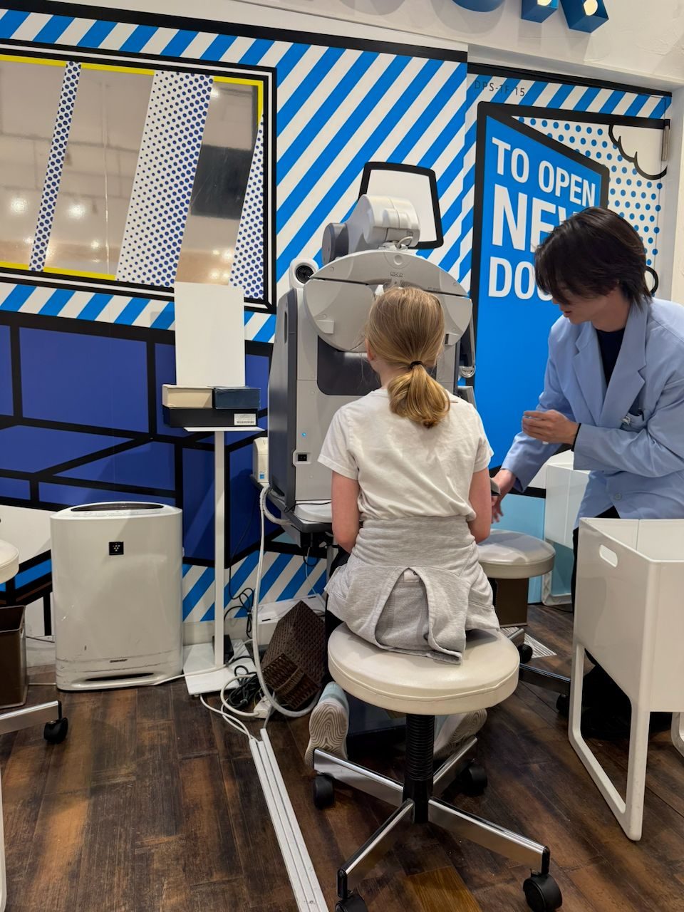 A young blonde girl sits at an eye examination machine while a staff member in a light blue coat attends to her, inside a boldly decorated optometrist's shop with blue and white graphic walls in Osaka.