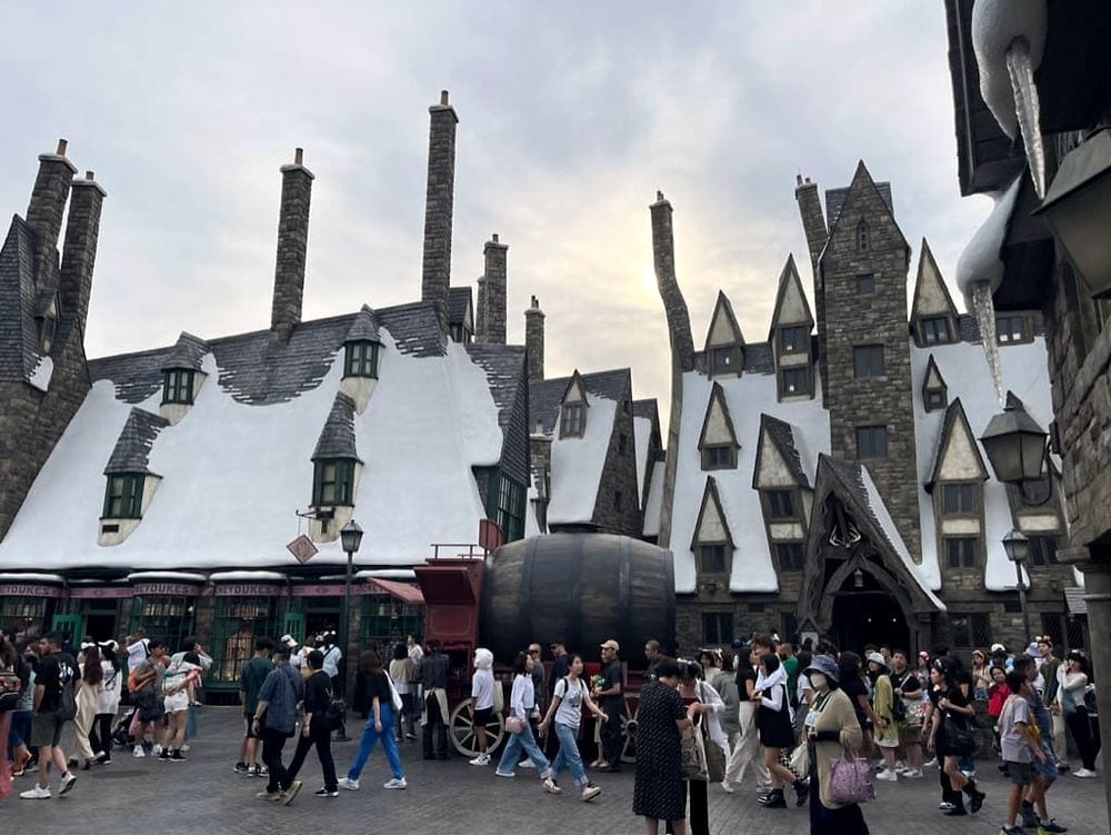 A wide shot of the Hogsmeade village area at The Wizarding World of Harry Potter in Universal Studios Japan, with snow-dusted peaked rooftops, a large barrel on a cart, and a crowd of visitors walking through the square under an overcast sky.