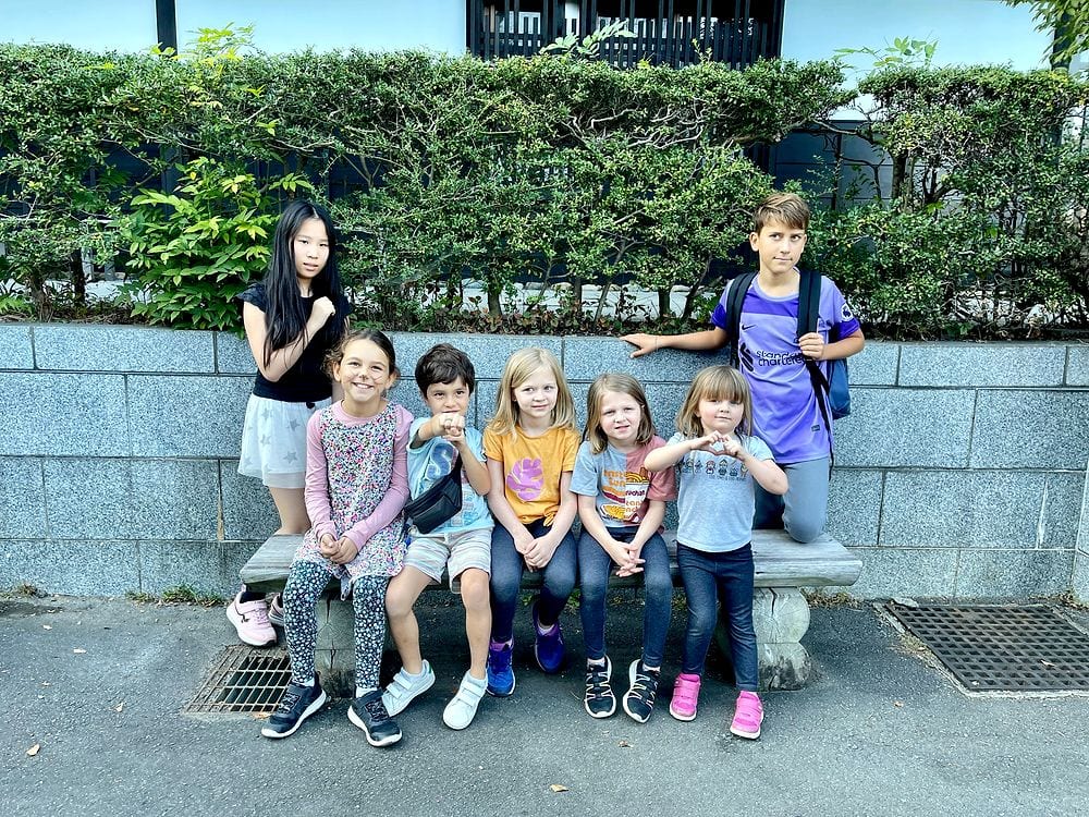 Seven children of varying ages pose together on a stone bench in front of a trimmed hedge wall in a residential area of Kawasaki, Japan, several making fist gestures toward the camera.