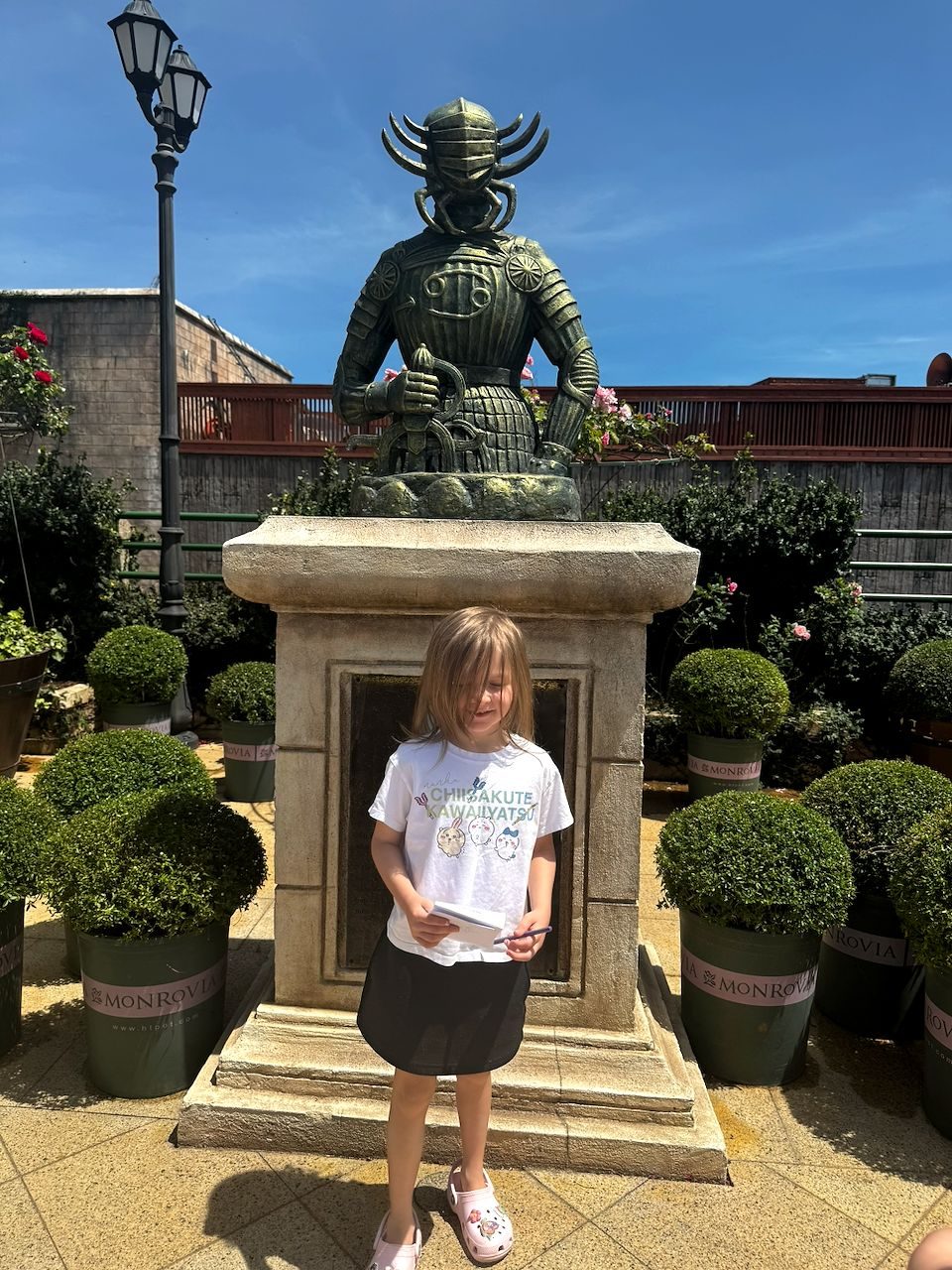 A young girl stands in front of a large bronze samurai statue on a stone pedestal, looking down at a notebook in her hands, surrounded by trimmed topiary shrubs under a blue sky.