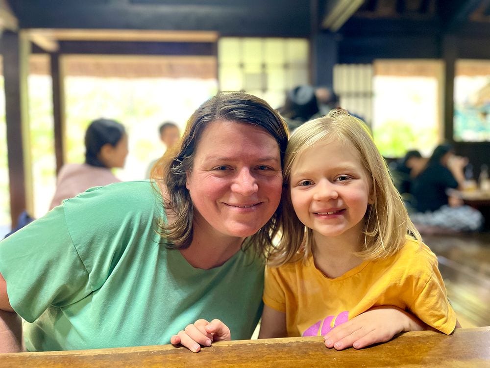 A woman in a green t-shirt and a young blonde girl in a yellow t-shirt smile together at a wooden table inside a traditional Japanese restaurant, with other diners visible in the softly lit background.