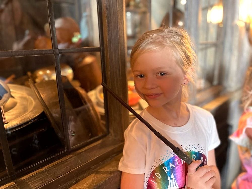 A young blonde girl holding a magic wand and smiling slightly at the camera inside a dimly lit wooden shop filled with brass props, at the Harry Potter area of Universal Studios Japan.