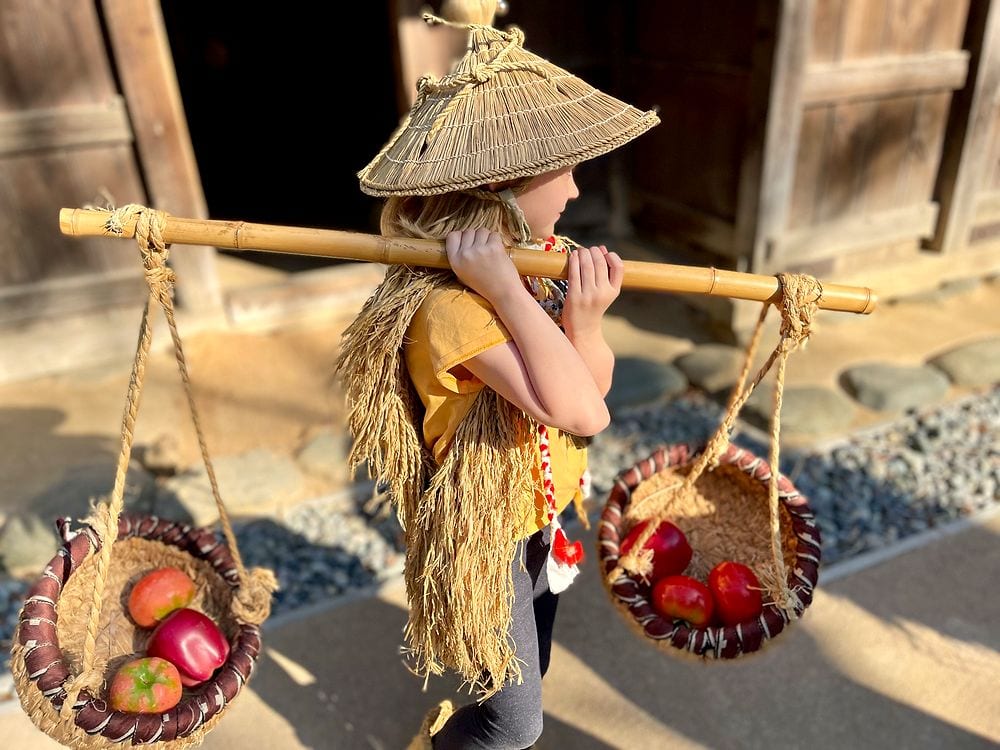 A young girl in a straw hat and cape carries a wooden yoke with two baskets across her shoulders in front of a historic thatched-roof farmhouse at Nihon Minkaen in Kawasaki.