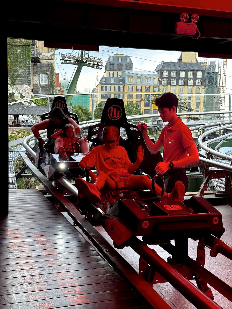 Two riders in race-car-style coaster seats are assisted by a staff member at the Ba Na Hills alpine coaster loading station, with the gondola cables and French-style buildings visible through the open-air gap behind them.