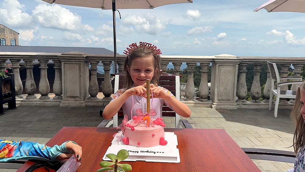 A young girl wearing a glittery Happy Birthday crown sits at an outdoor table on a stone terrace, hands gripping a tall birthday candle on a pink heart-decorated cake, with a blue sky and mountain views behind her.