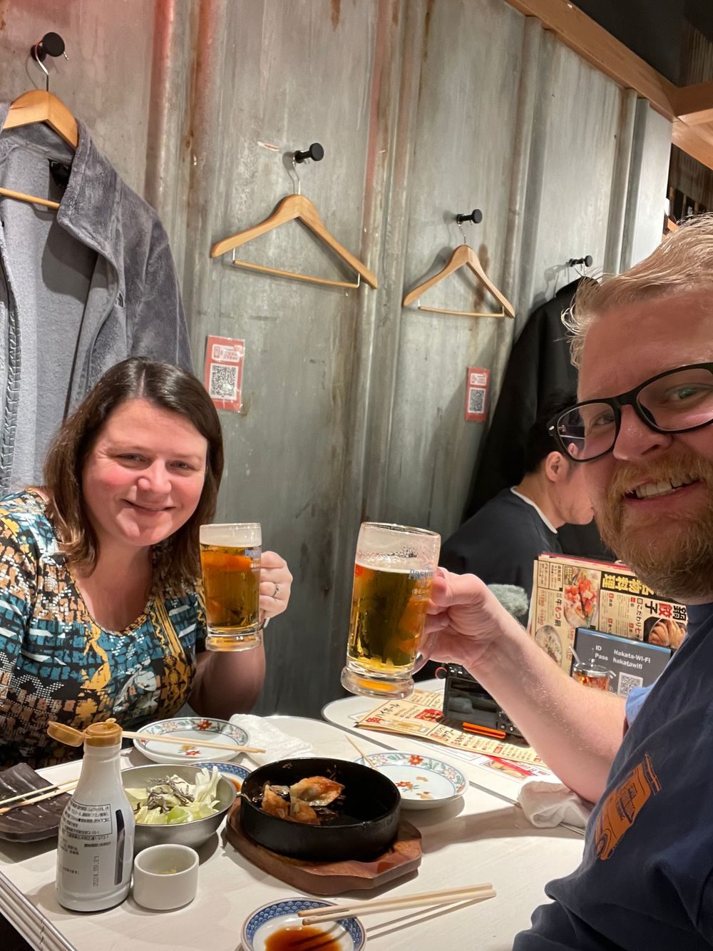 Two people raise beer glasses at a small table inside a Golden Gai bar, with coats hanging on hooks along the wall behind them.