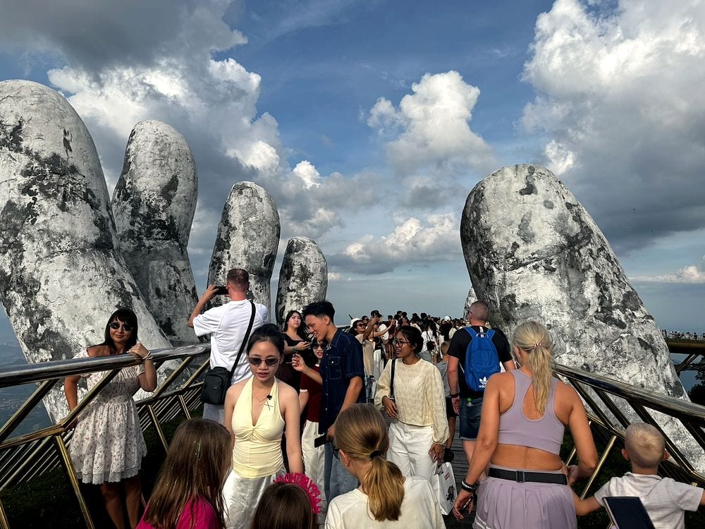 A dense crowd of tourists walking along the Golden Bridge at Ba Na Hills between the two giant stone hands under a partly cloudy sky