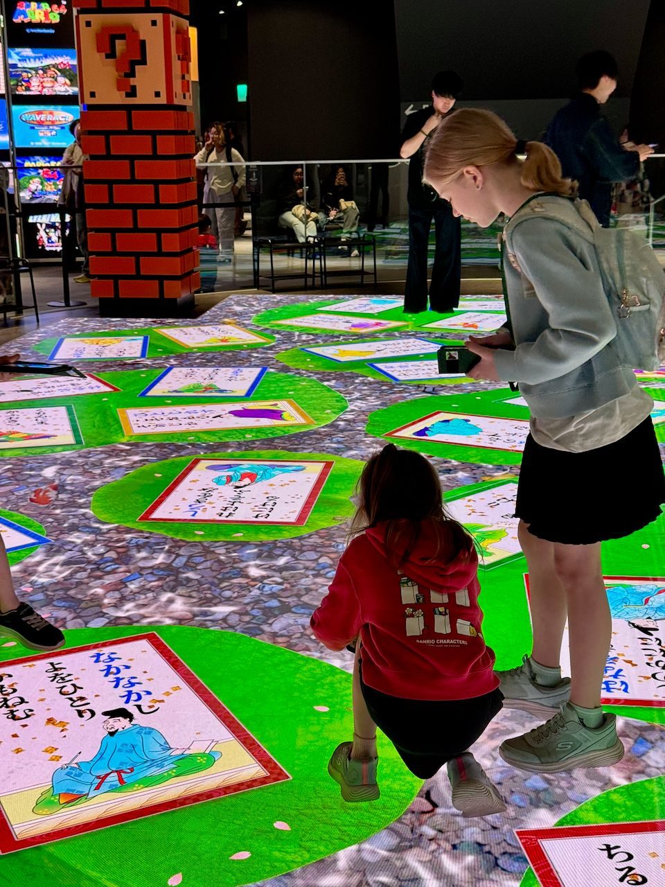 Two girls crouch and stand over a large illuminated floor projection displaying colorful karuta poetry cards with Japanese text and illustrations, inside a museum with a Super Mario question block display in the background.