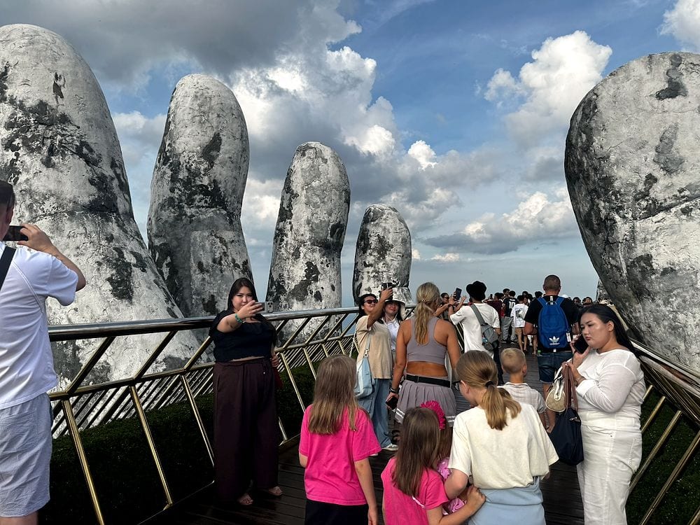 A dense crowd of tourists walking along the narrow Golden Bridge walkway at Ba Na Hills, flanked by the large stone hand sculptures under a moody sky