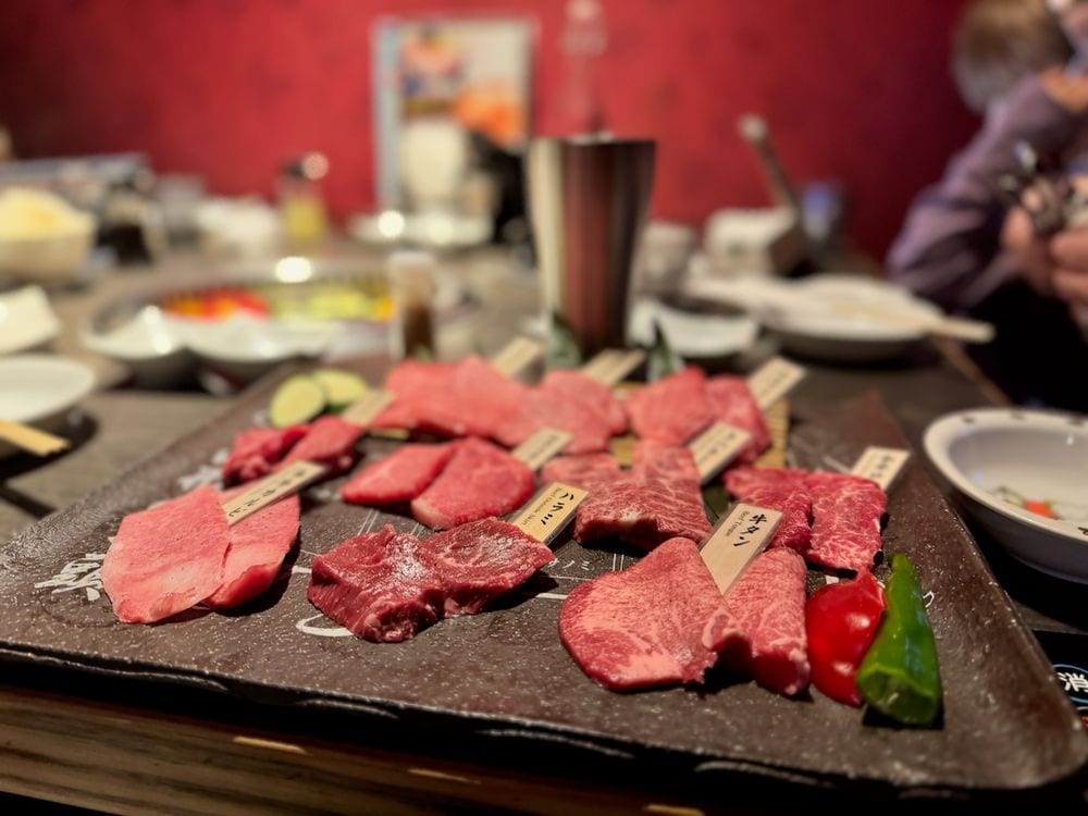 A slate platter of labeled raw wagyu beef cuts arranged for yakiniku grilling, with small wooden tags identifying each cut, set on a restaurant table with side dishes and drinks blurred in the background.