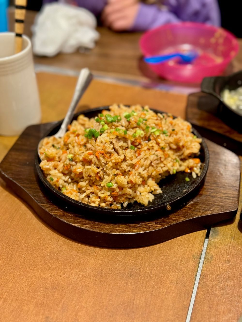 A sizzling plate of garlic fried rice with green onion on a wooden-handled iron skillet, served at a restaurant table.