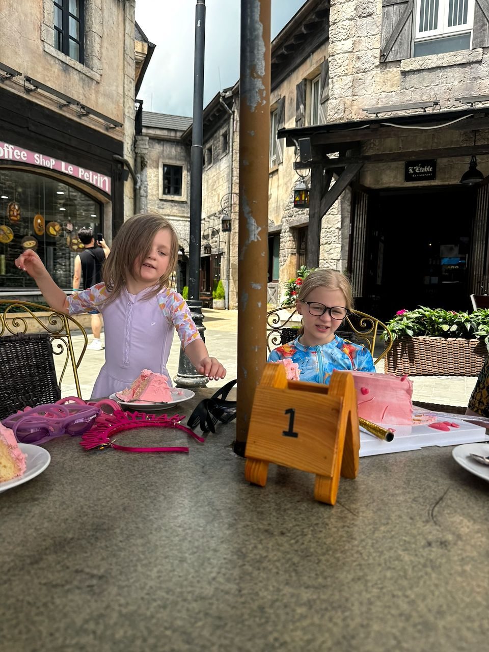 Two young girls sit at an outdoor table in a French village-style courtyard at Ba Na Hills, with slices of pink birthday cake in front of them and a pink birthday crown on the table.