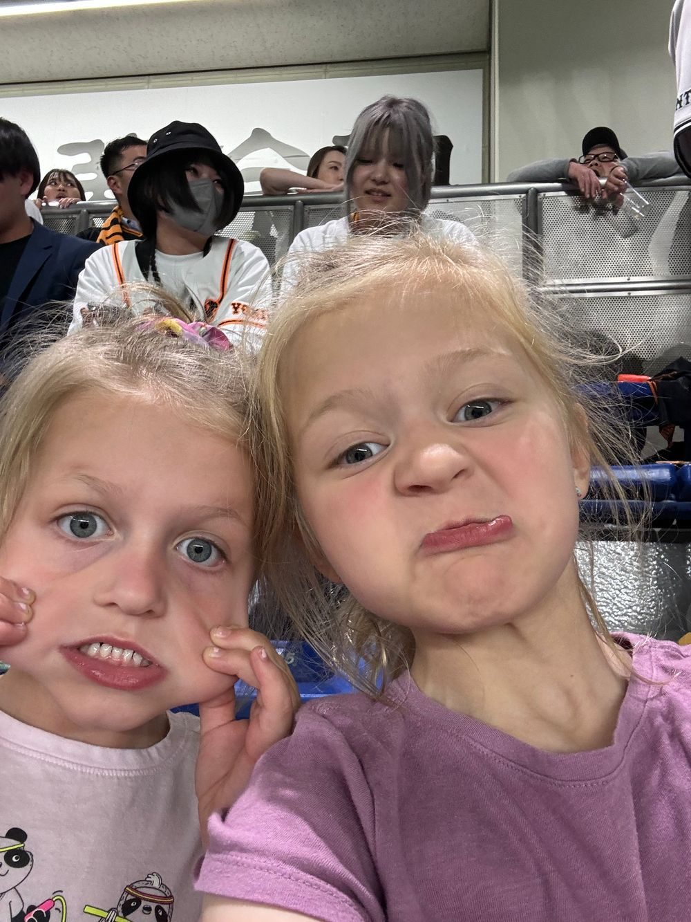 Two young girls make exaggerated grimacing faces for a selfie at Tokyo Dome, with fans visible in the stands behind them.