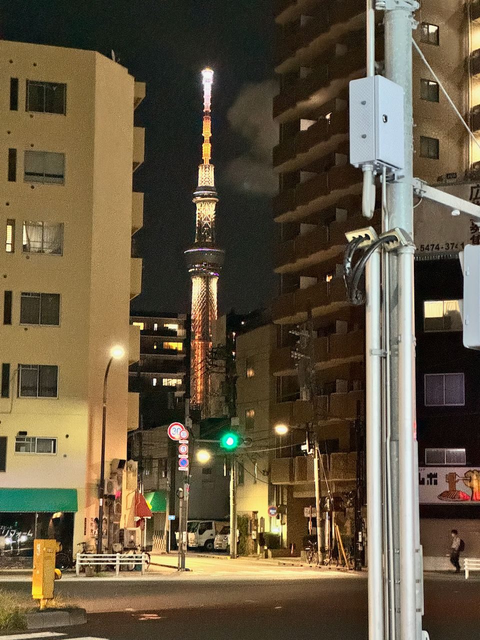 Tokyo Skytree glowing orange against a dark night sky, framed at the end of a quiet residential street in Taito with traffic signals and low-rise apartment buildings on either side.