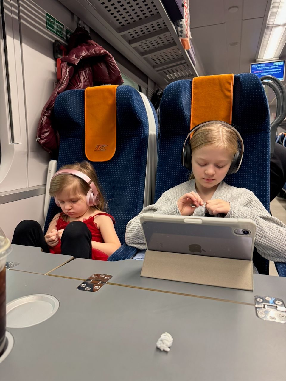 Harper and Lily sitting side by side on a Polish intercity train wearing headphones, with an iPad propped on the tray table