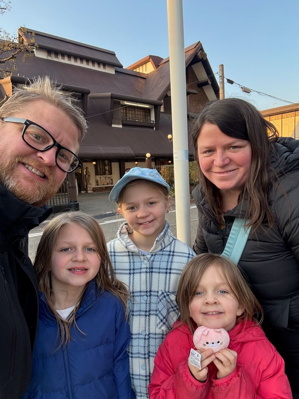 A family of five takes a selfie outside a traditional Japanese building in winter light, with two adults and three young girls, the youngest holding a pink stuffed animal.