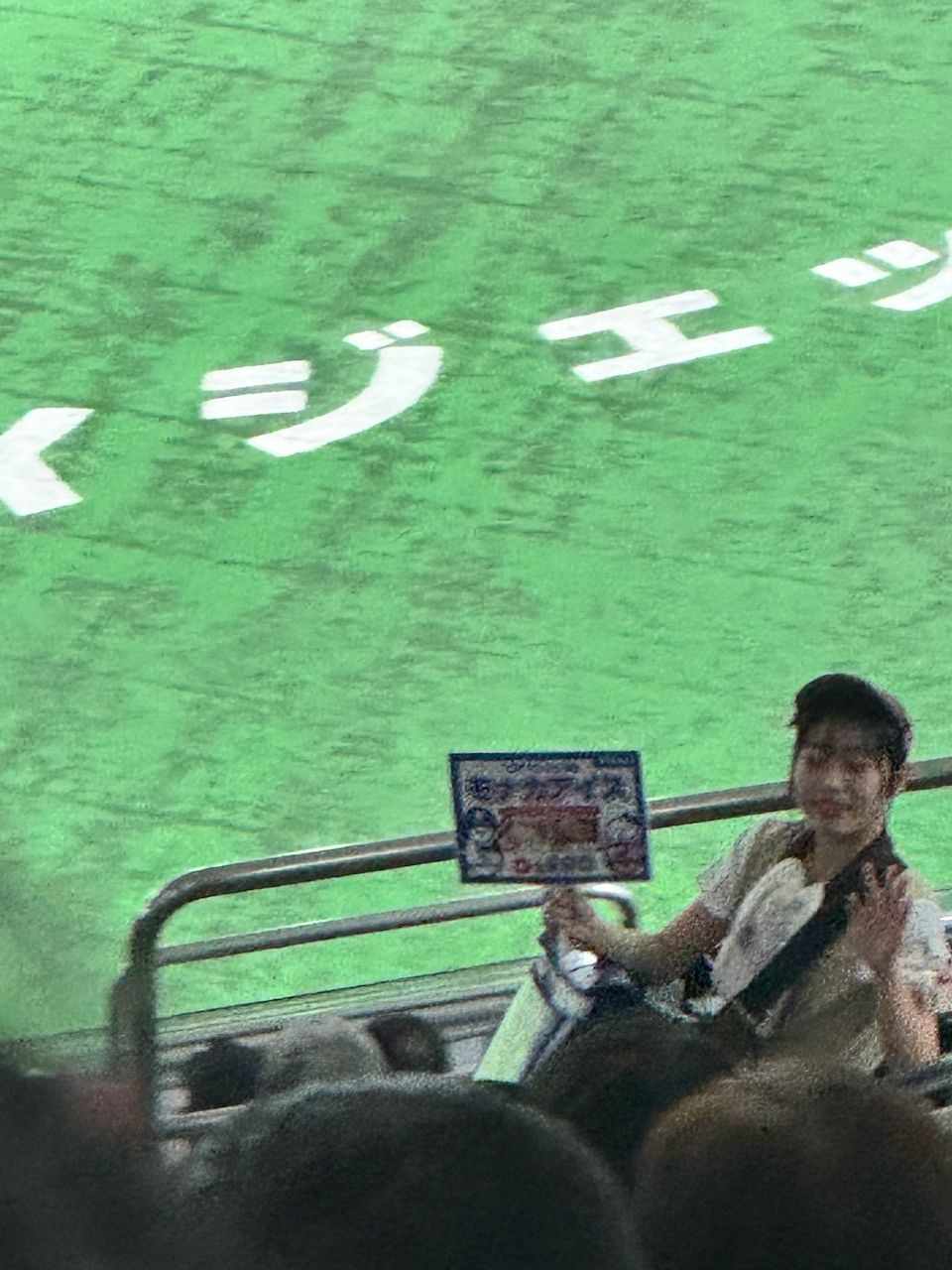 A stadium vendor at Tokyo Dome holds a menu card while seated at the aisle railing, with the illuminated green field and Japanese katakana lettering visible in the background.