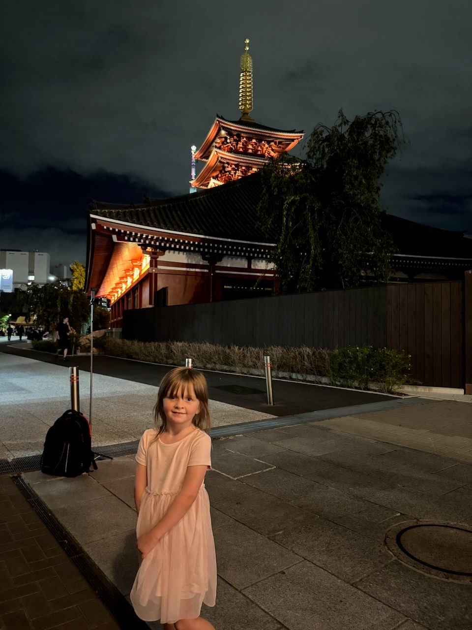 A young girl in a light pink dress stands on a paved path at night in front of the illuminated pagoda and lantern-lined gate of Senso-ji temple in Asakusa, with the Tokyo Skytree faintly visible in the background.