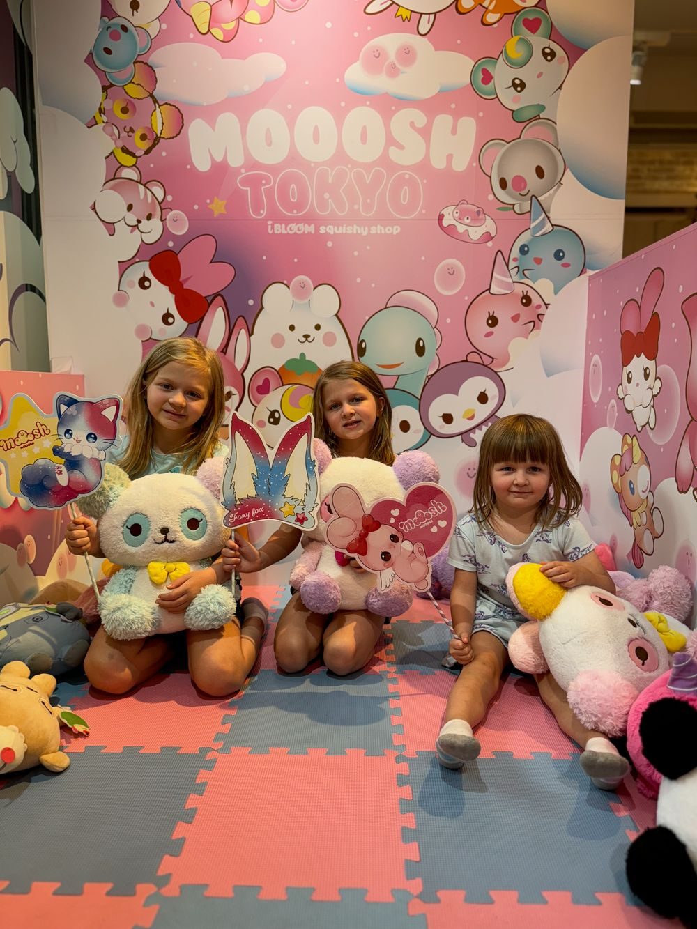 Three young girls kneel together holding large squishy plush toys inside MOOOSH TOKYO, a pink character-themed store in Harajuku, surrounded by shelves of squishy characters.