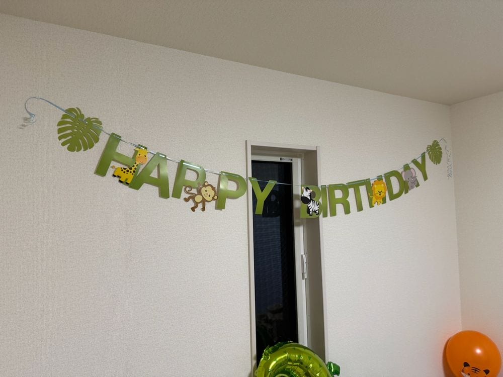 A green jungle-themed Happy Birthday banner with cartoon animals including a giraffe, monkey, zebra, and lion strung across a white apartment wall, with orange and green balloons visible in the corner.