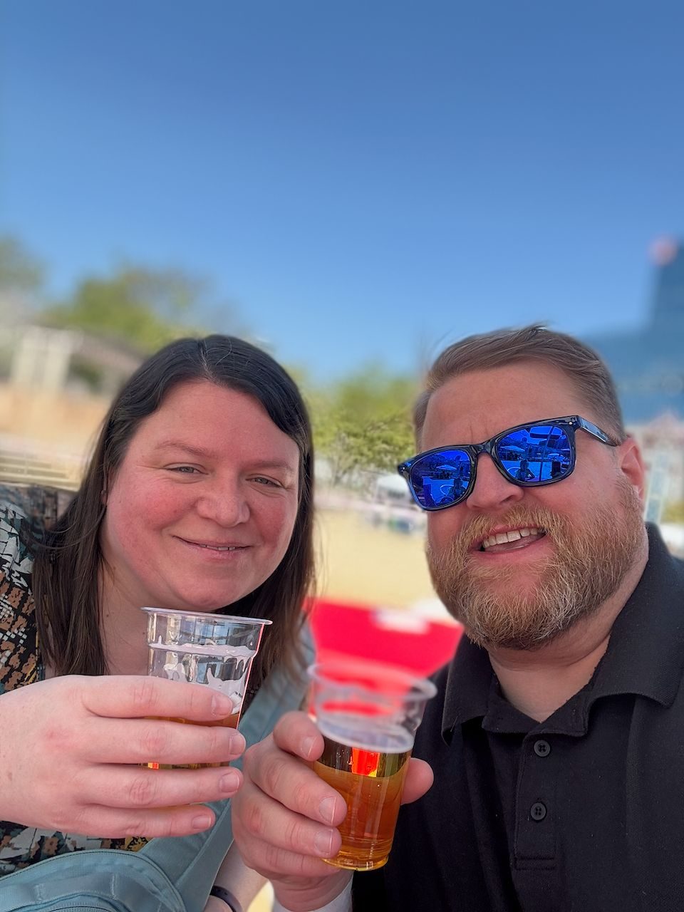 Adam and Lindsay smile at the camera holding plastic cups of beer outdoors under a clear blue sky at Ogimachi Park in Osaka.