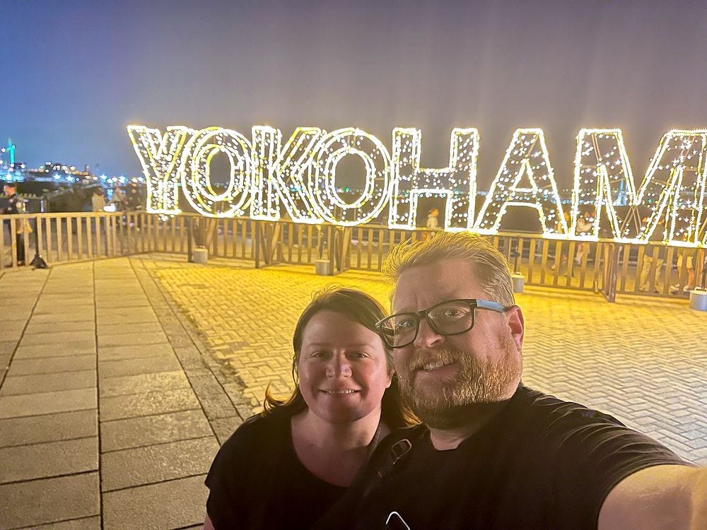 A man and woman take a selfie in front of a large illuminated YOKOHAMA sign at night by the waterfront.