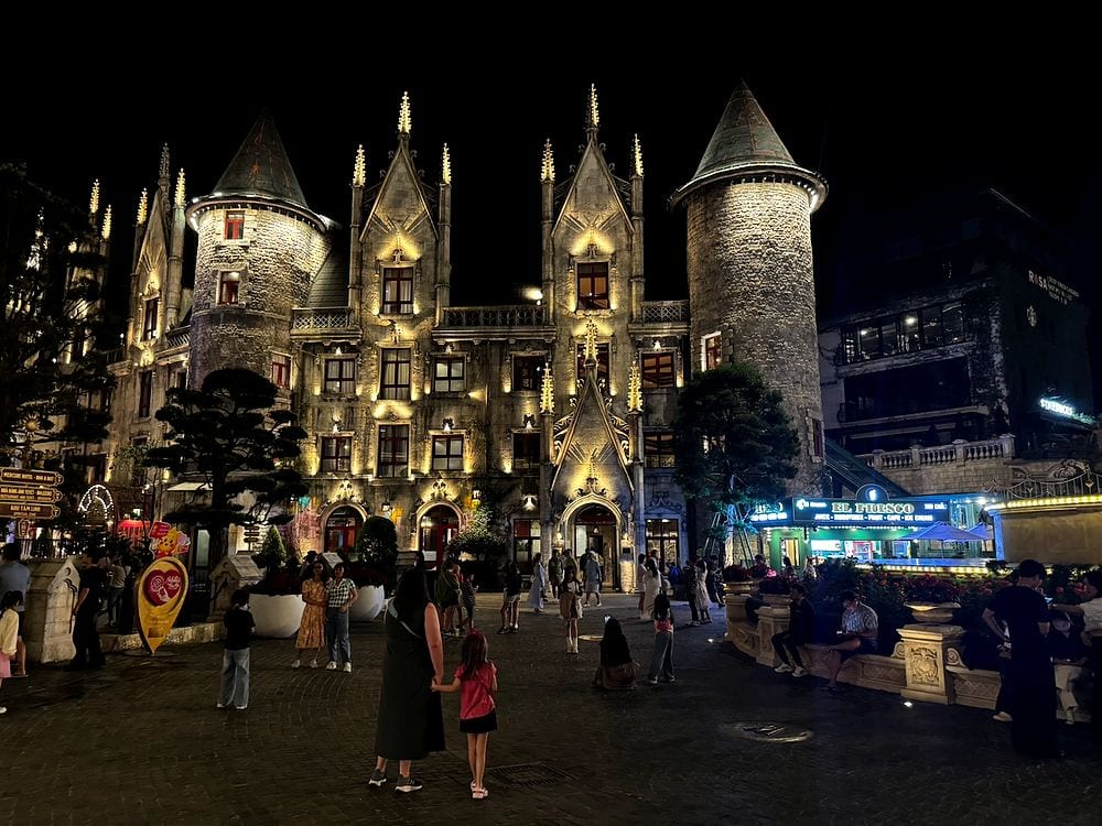 An illuminated Gothic-style stone castle with pointed towers glowing against a black night sky at Ba Na Hills, with a crowd of visitors walking through the plaza below