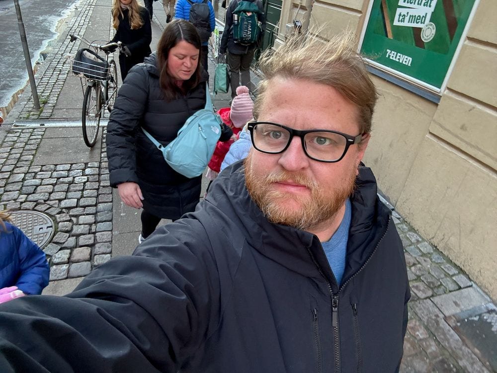 A man takes a windswept selfie on a Copenhagen sidewalk while a woman walks behind him past a 7-Eleven storefront