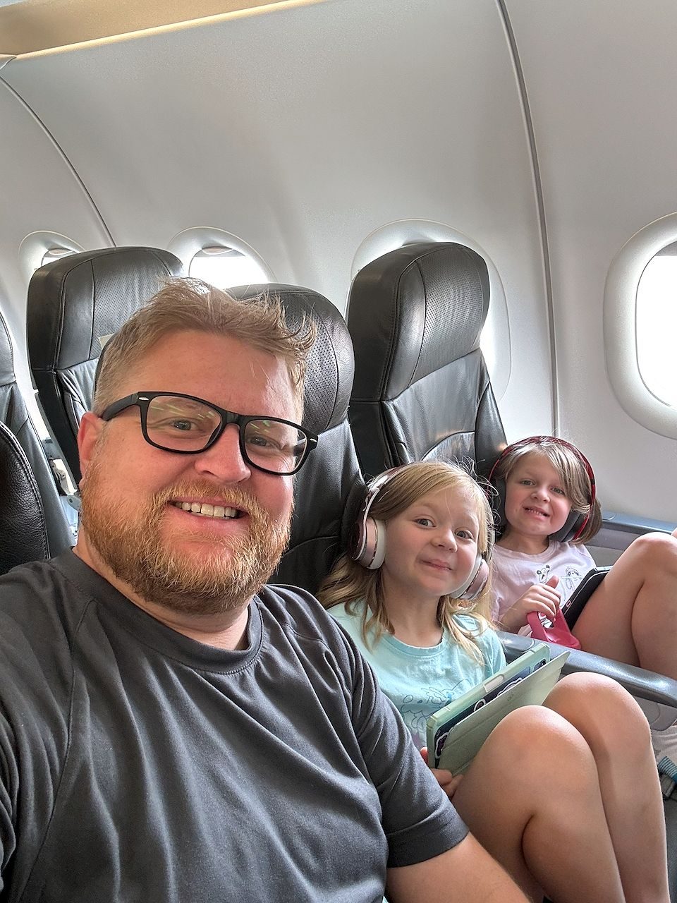 A man takes a selfie on an airplane with two young girls seated behind him, both wearing headphones and holding tablets, with cabin windows visible in the background.