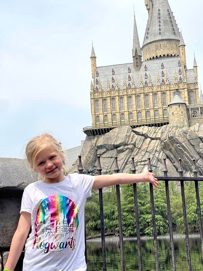 A young blonde girl in a rainbow Hogwarts t-shirt smiles while leaning on a black iron railing, with the Hogwarts Castle replica at Universal Studios Japan rising behind her.