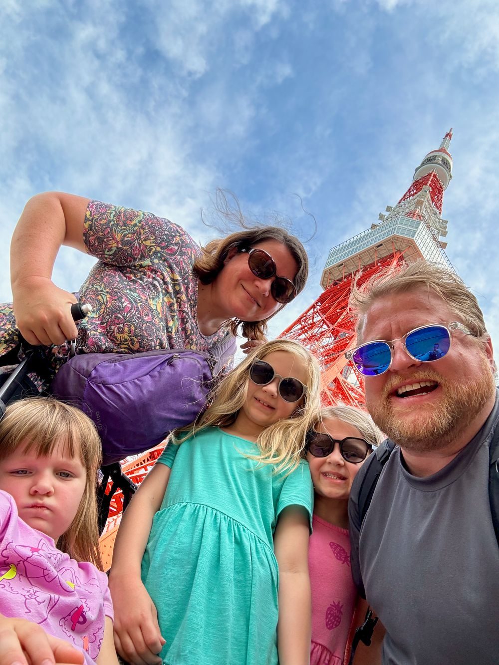 A low-angle family selfie with Adam, Lindsay, and their three daughters in front of Tokyo Tower on a partly cloudy day, with the red lattice tower rising into the sky behind them.