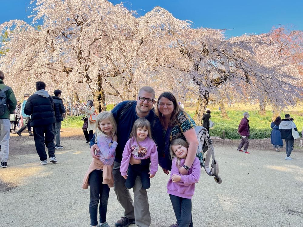 A family of five, two adults and three young girls, pose smiling in front of large weeping cherry blossom trees in full bloom at a crowded park on a sunny day.