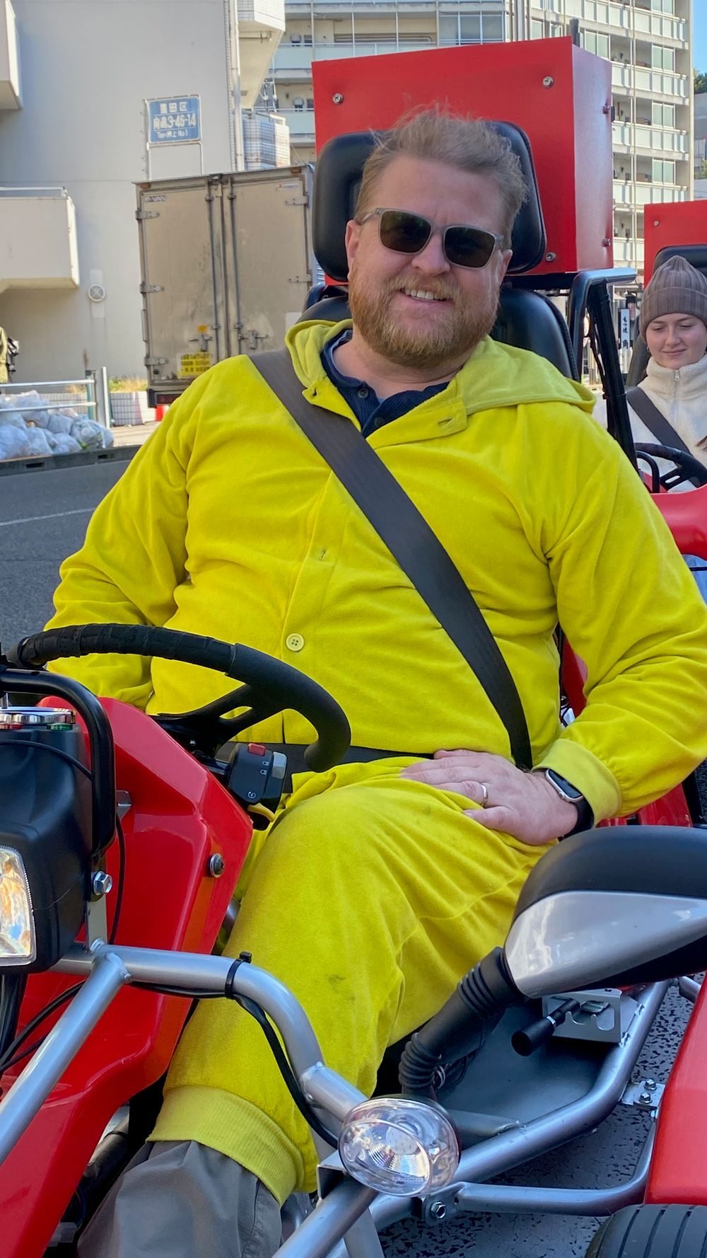 Adam sits in a red street kart wearing a bright yellow Pikachu onesie and sunglasses, seatbelt on, smiling at the camera with Tokyo streets visible behind him.