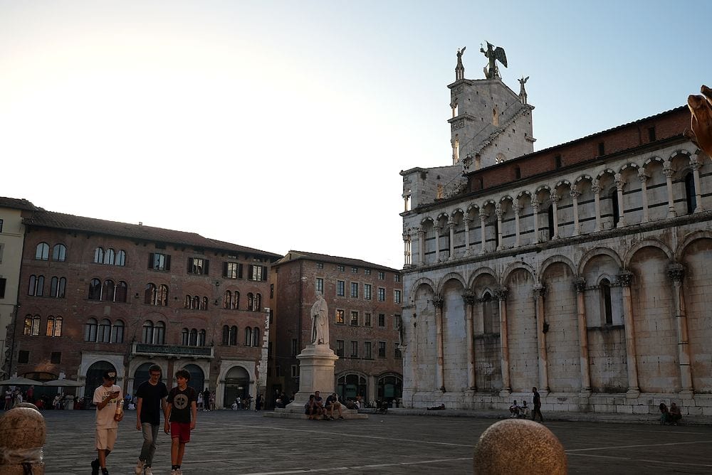 Wide shot of Piazza San Michele in Lucca at dusk, with the marble facade of San Michele in Foro church on the right and warm evening light across the square