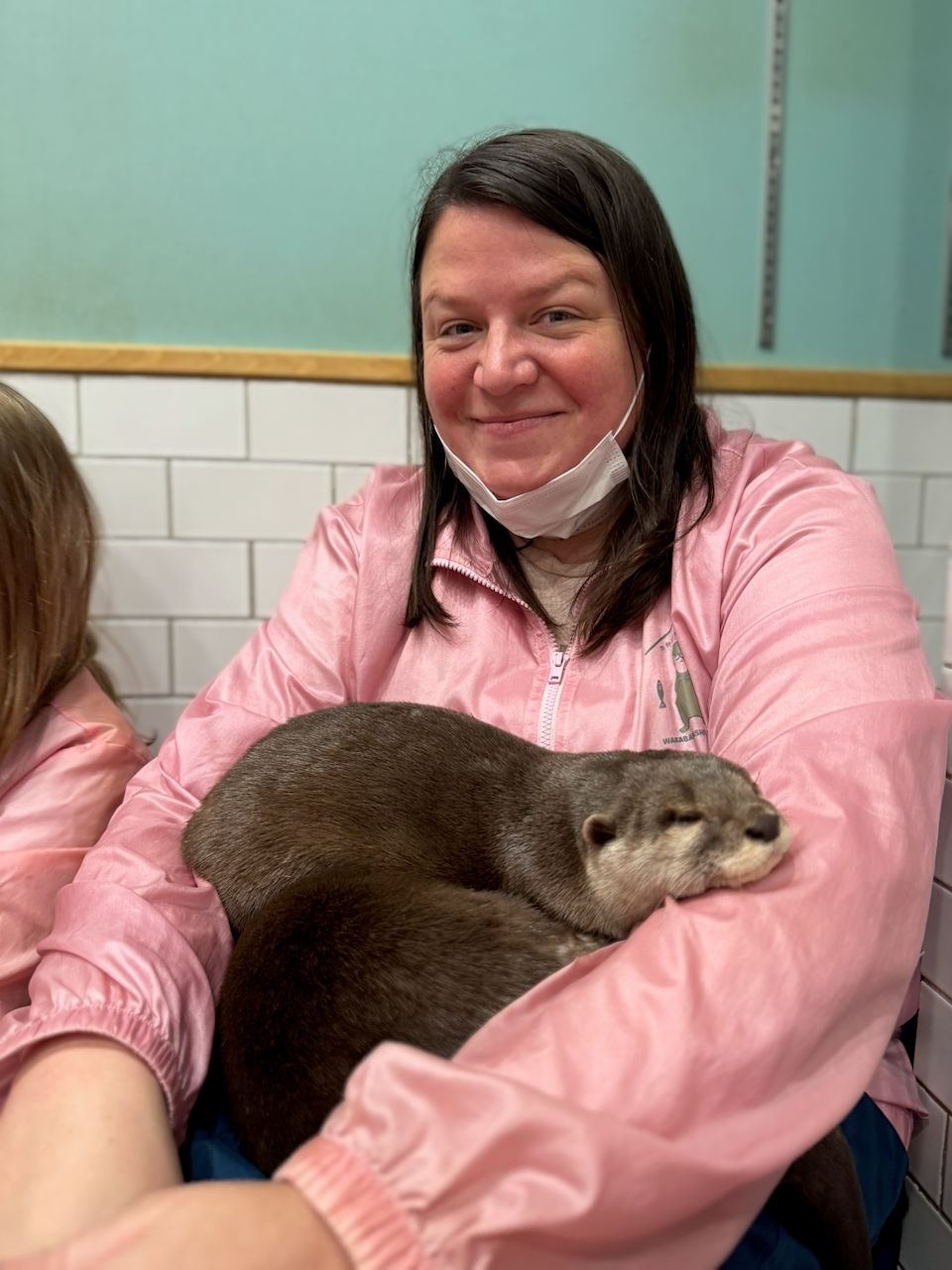 A woman in a pink jacket holds a small sleeping otter cradled in her arms inside a tiled animal cafe in Shibuya, Japan.