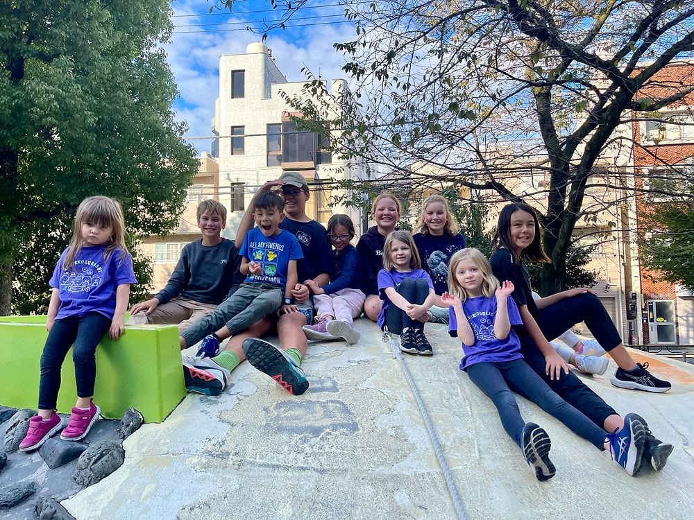 A group of about ten children sitting and posing on a large curved concrete slide structure in a small urban park in Tokyo, several wearing purple shirts, with trees and city buildings visible behind them.