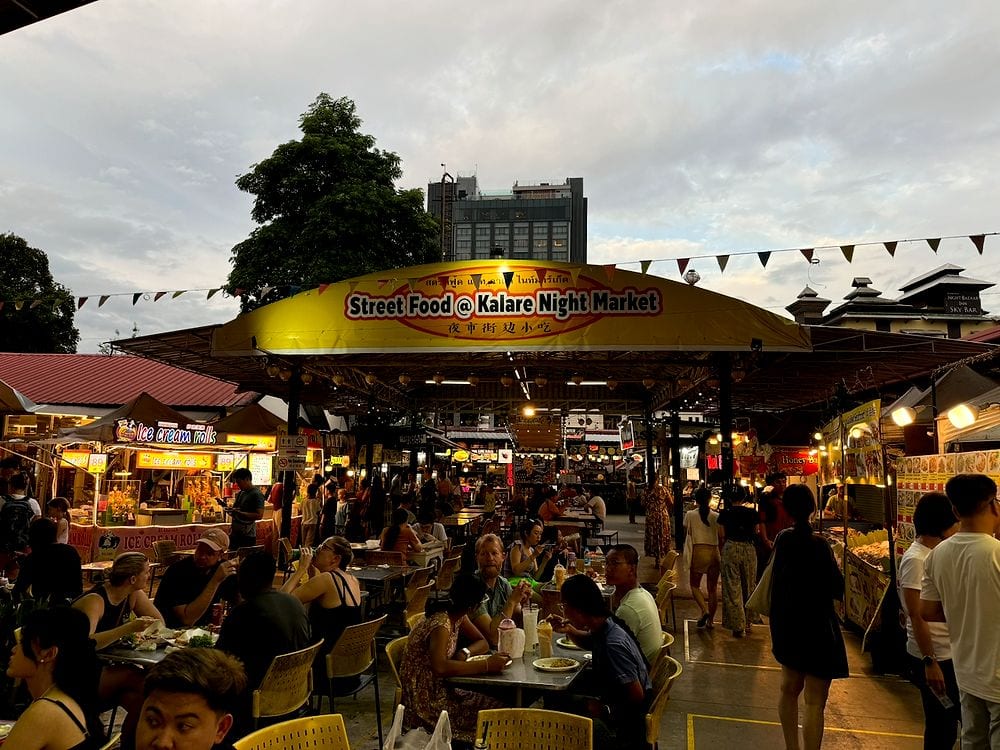 A wide shot of the Kalare Night Market in Chiang Mai at dusk, with a large yellow sign reading Street Food at Kalare Night Market, crowded tables of diners, surrounding food stalls lit up, and an overcast evening sky above.