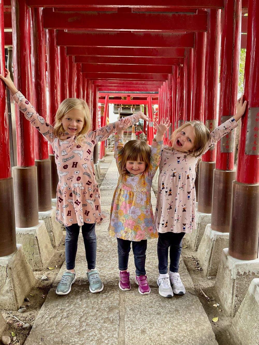 Lily, Cora, and Harper stand in a row with arms raised inside a tunnel of red torii gates at a Shinjuku shrine, all three smiling at the camera.