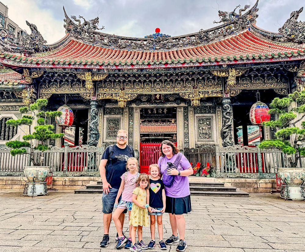 A family of five stands in front of Longshan Temple in Taipei, with its ornate black and gold facade, red roof, dragon carvings, and red paper lanterns visible behind them.