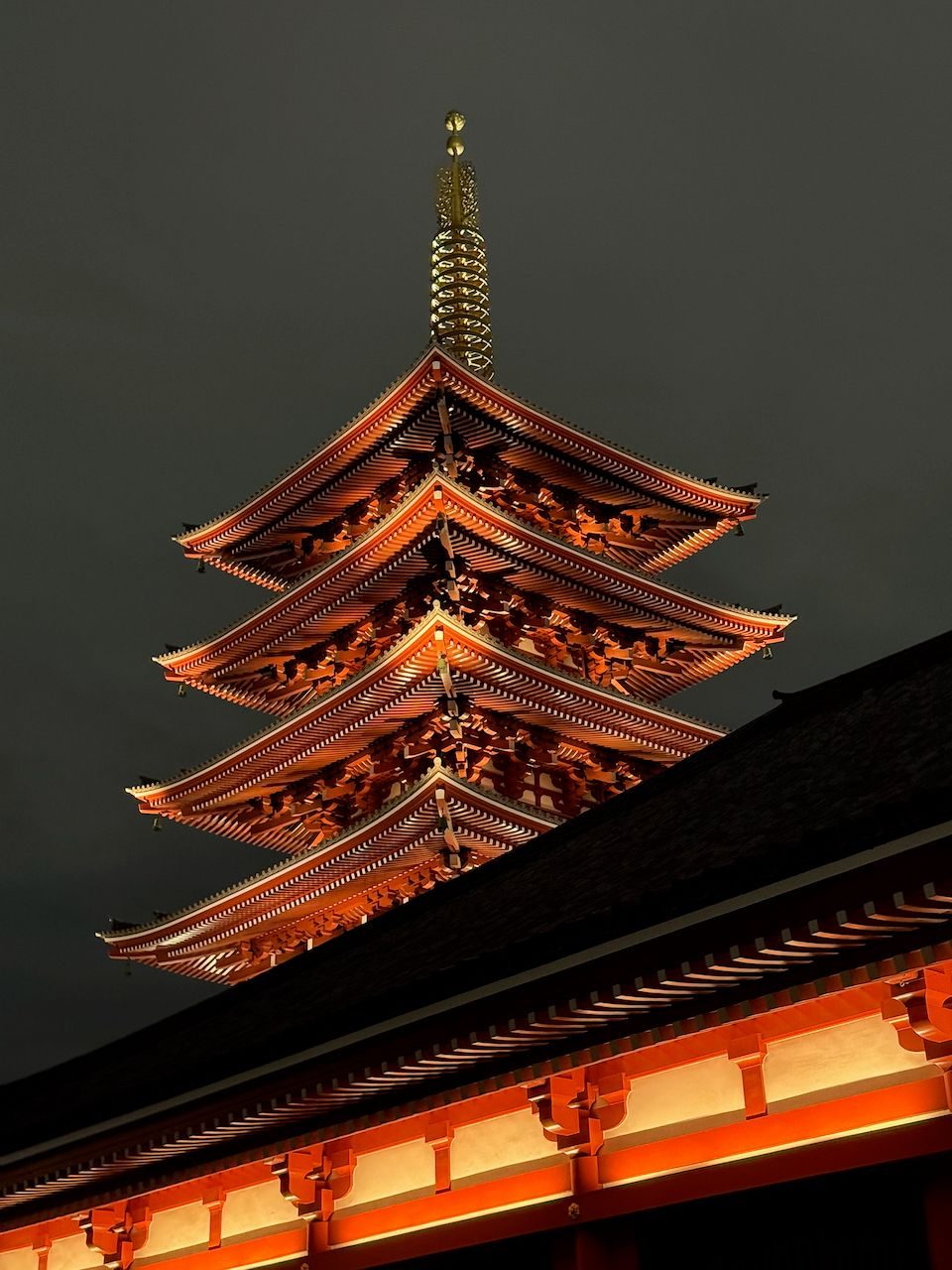 The illuminated five-story pagoda of Senso-ji temple rises against a dark night sky in Asakusa, its orange-red tiers glowing warmly against the darkness.