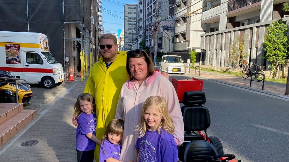 A family of five poses on a Tokyo street, the father wearing a yellow Pikachu costume and the mother in a pink onesie, with three young girls in matching purple shirts standing in front of them, a red street kart visible to the right.