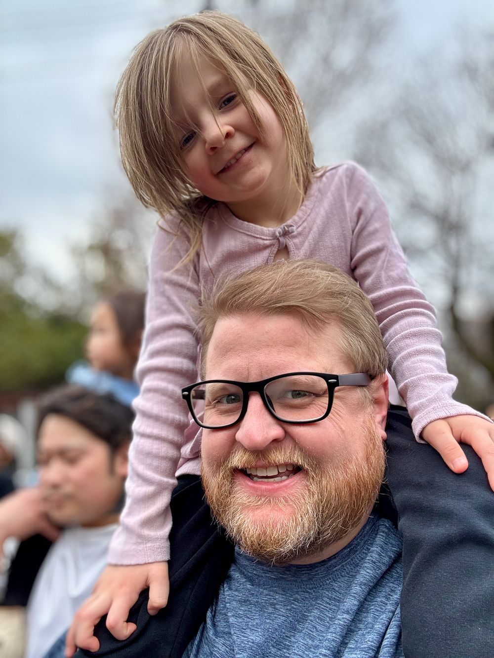 A young girl with blonde hair sits on a bearded man's shoulders, both smiling at the camera outdoors at Tennoji Zoo in Osaka, Japan.