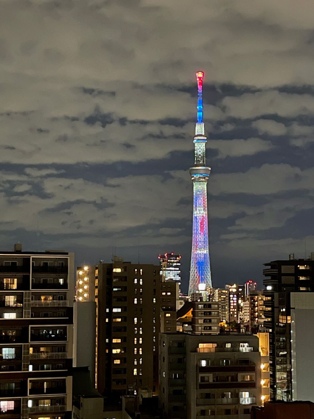 Tokyo Skytree lit up at night in a colorful illumination pattern, rising above a dense cityscape of illuminated apartment buildings.