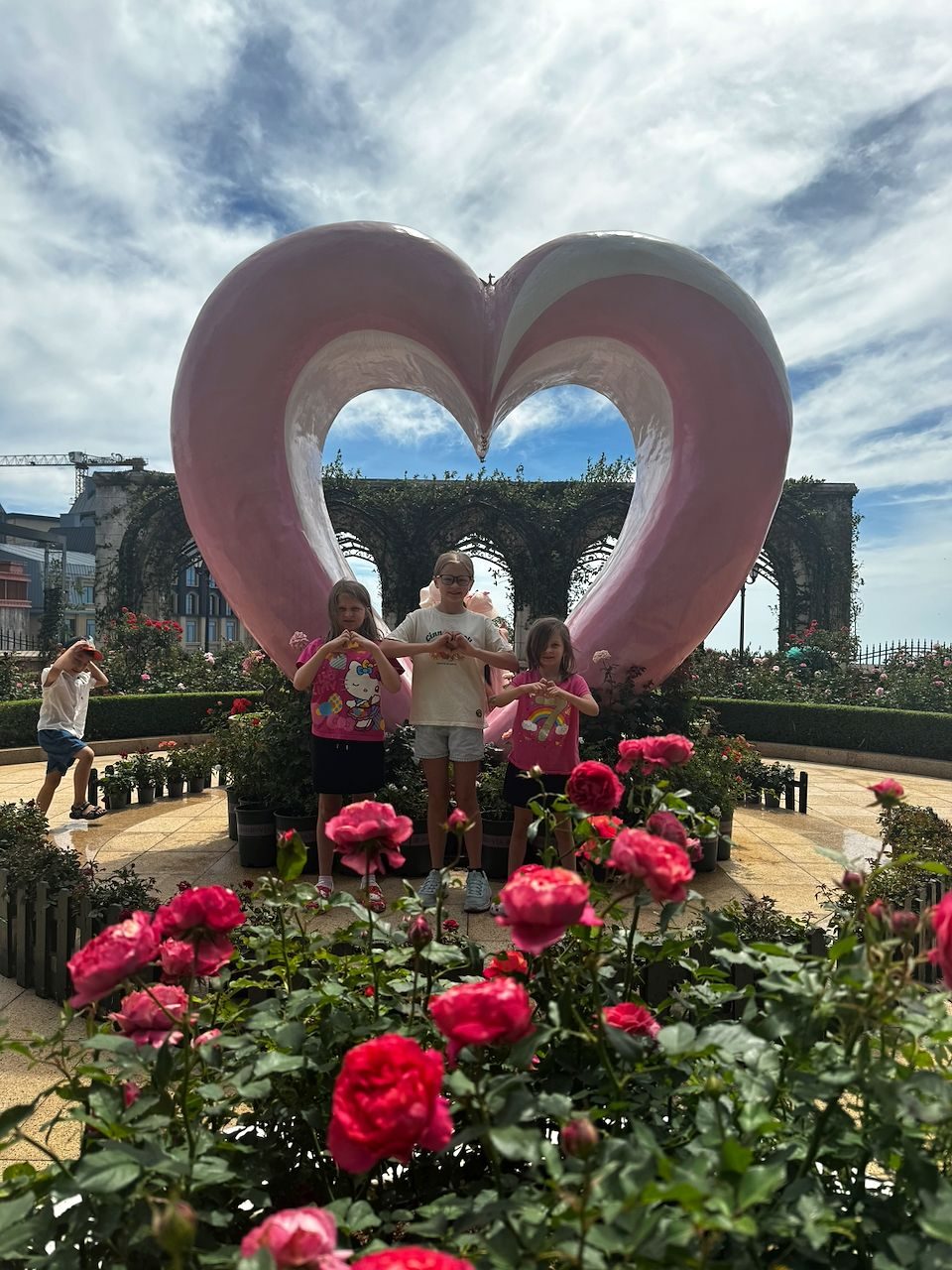 Three girls stand in front of a large pink heart sculpture surrounded by pink roses in a formal garden, making heart shapes with their hands, with ivy-covered archways and a partly cloudy sky behind them.