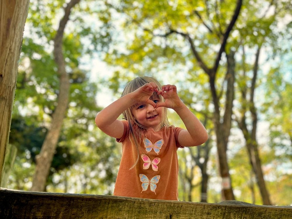 A young girl makes a heart shape with her hands in front of a wooden play structure at Heiwa no Mori Park, with trees visible in the background.