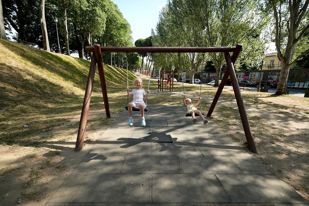 Two young girls sit on swings at an outdoor playground along the tree-lined path on top of the Lucca city walls on a sunny summer day