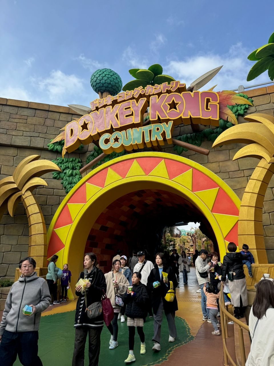 The entrance arch to Donkey Kong Country at Universal Studios Japan, with a large colorful sign in English and Japanese above a yellow and red sunburst arch, crowds of visitors walking through and around the entrance on a partly cloudy day.
