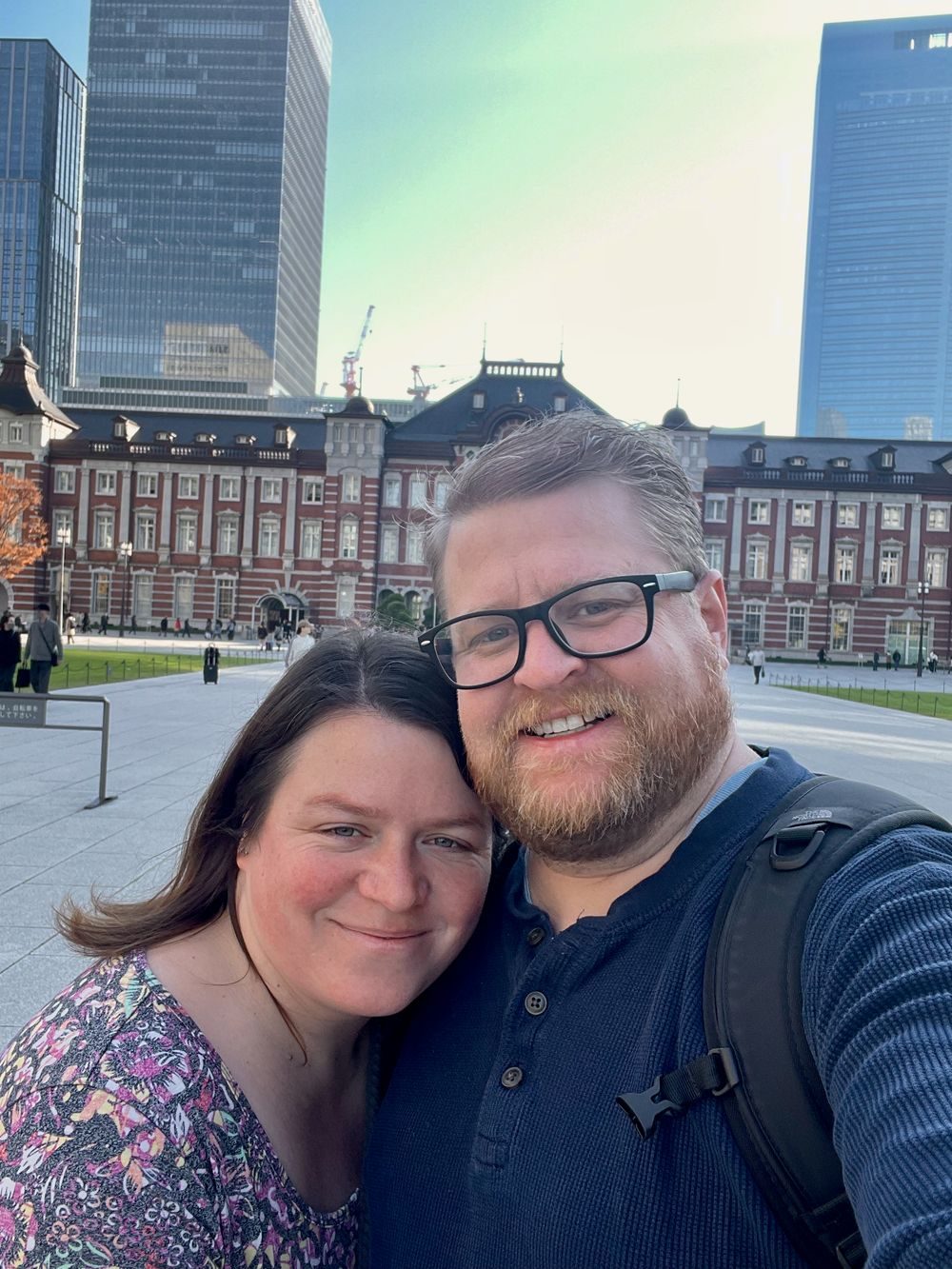 Adam and Lindsay take a selfie in front of the red brick Marunouchi facade of Tokyo Station, with modern office towers visible behind them.